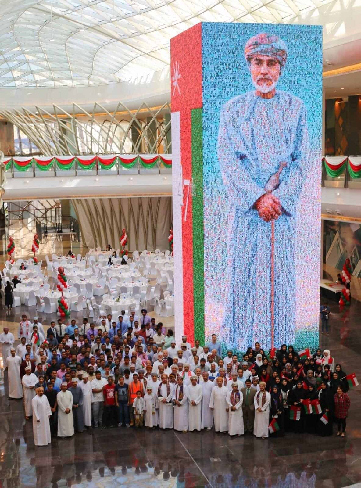 Four-sided LED tower in the atrium of Muscat Grand Mall showing a photo-mosaic portrait of the Sultan of Oman beside the Omani flag, with mall staff gathered in front for National Day