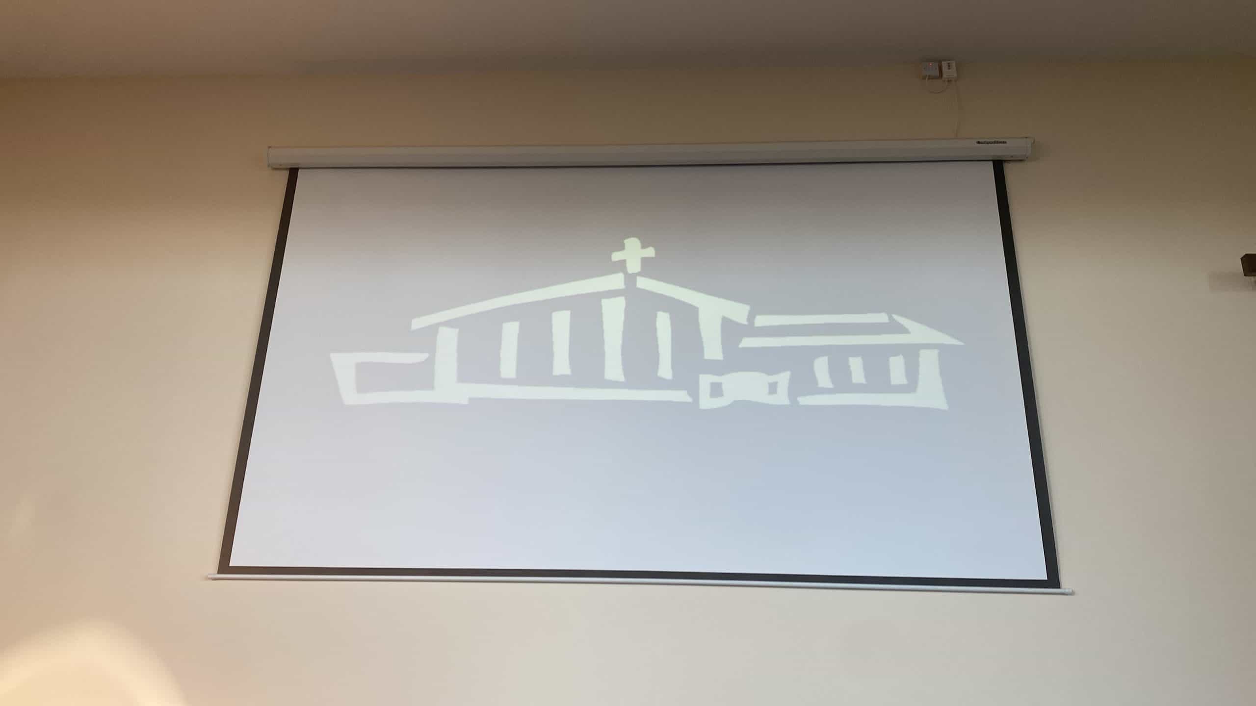 Retractable projector screen in a church hall showing a church logo washed out to pale grey by daylight from the windows