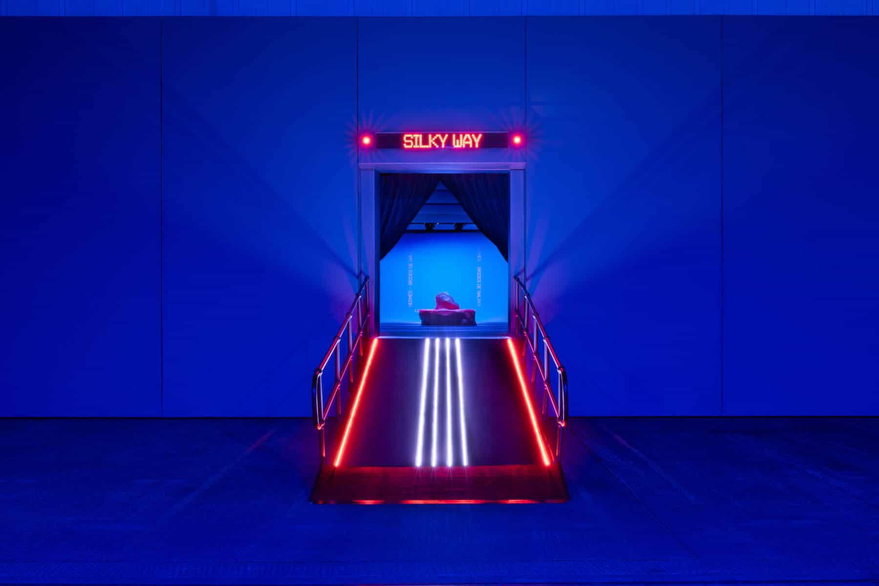 Red dot-matrix LED ticker reading SILKY WAY above the entrance doorway of the Hermès Brides de Galaxy event, in a blue-lit room with a neon-edged ramp