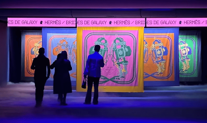 Long pink LED ticker scrolling HERMÈS / BRIDES DE GALAXY above a row of oversized Hermès silk scarf designs, with three guests in silhouette in front