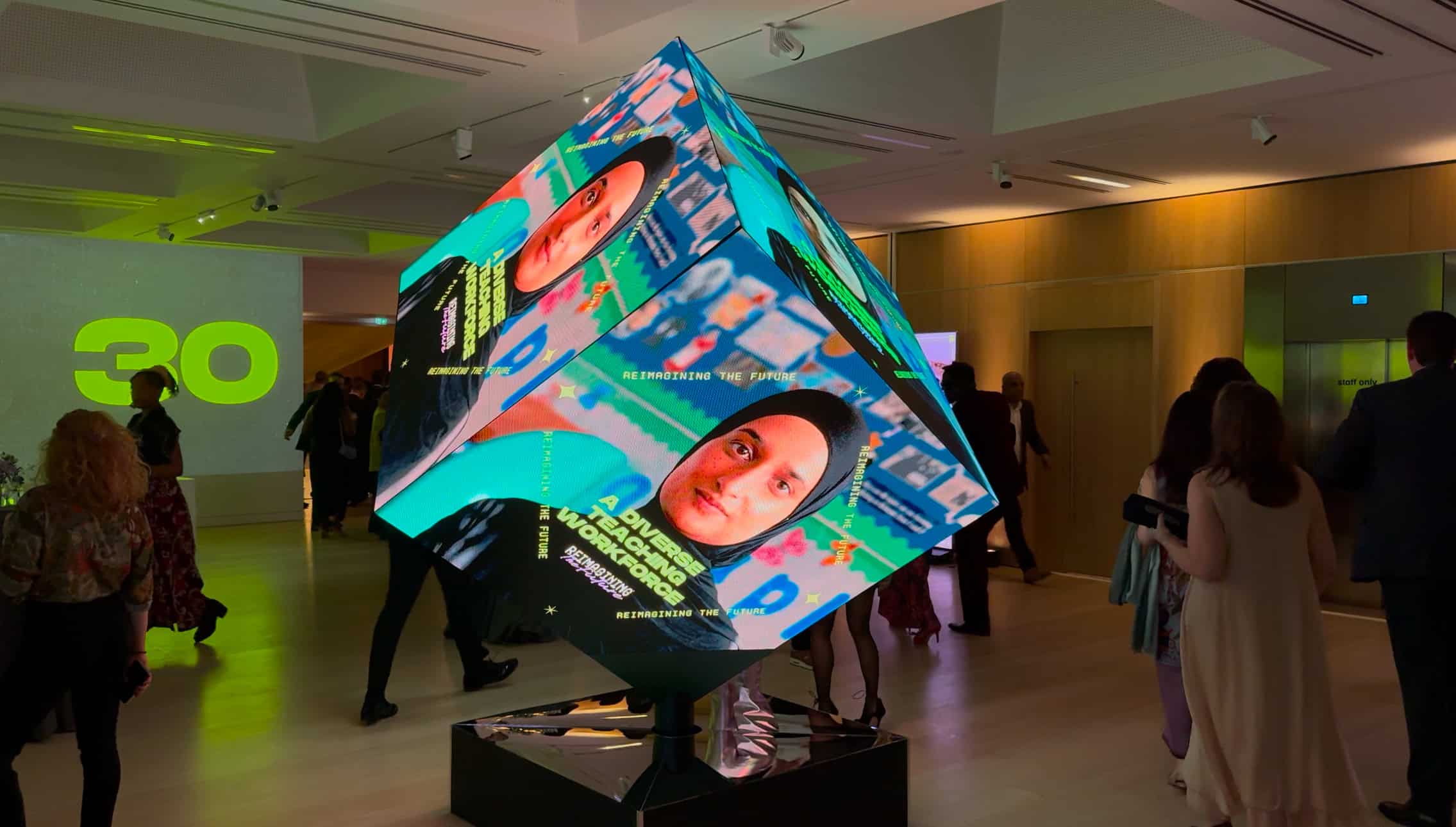 2.8 m LED cube standing on one corner on a mirrored plinth at the Mission 44 Reimagining the Future event, its faces showing a portrait and the words A Diverse Teaching Workforce as guests walk past