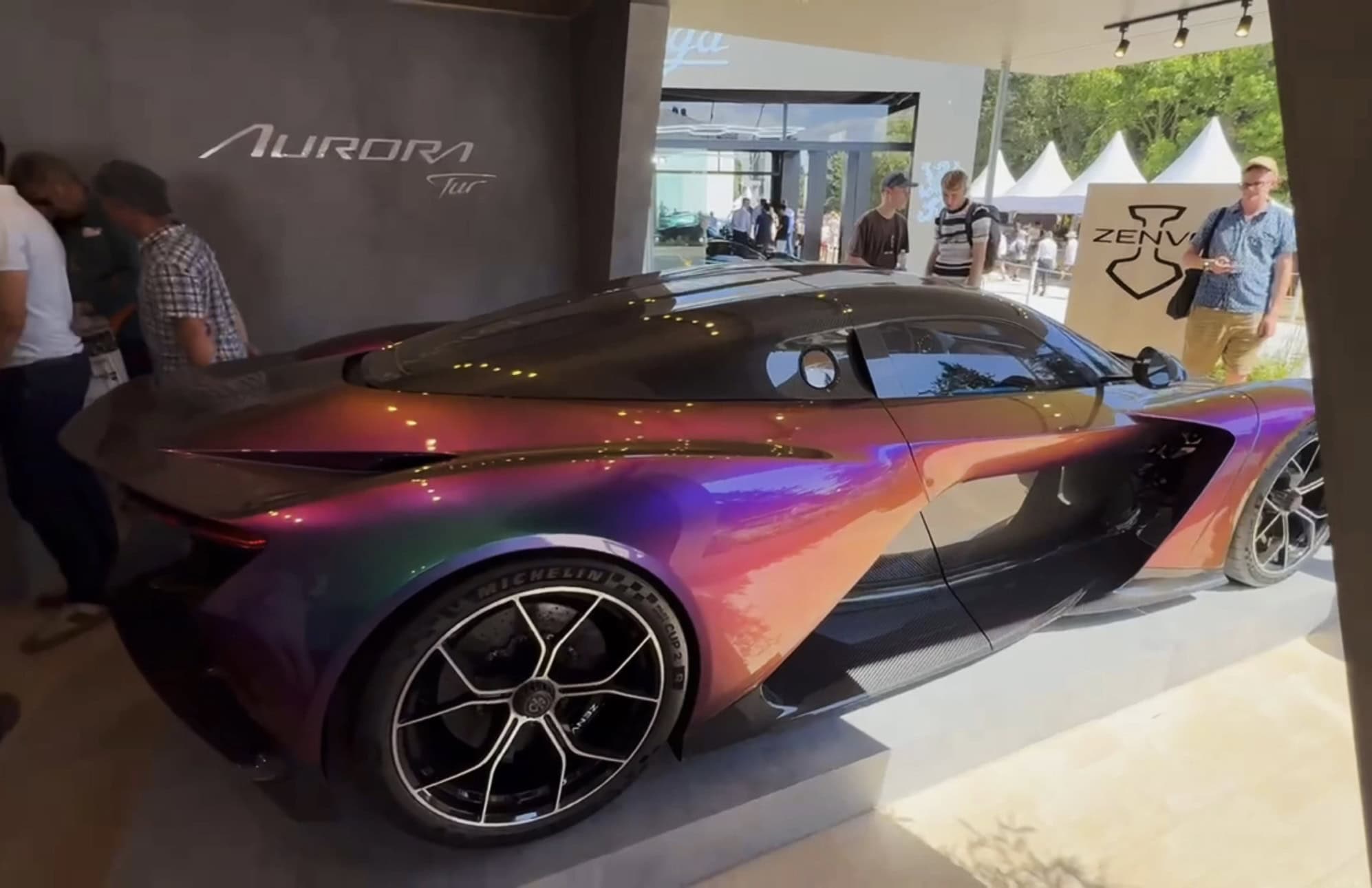 Side view of the iridescent purple and orange Zenvo Aurora Tur on its stand at the Goodwood Festival of Speed, Aurora Tur lettering on the grey wall behind and visitors looking on
