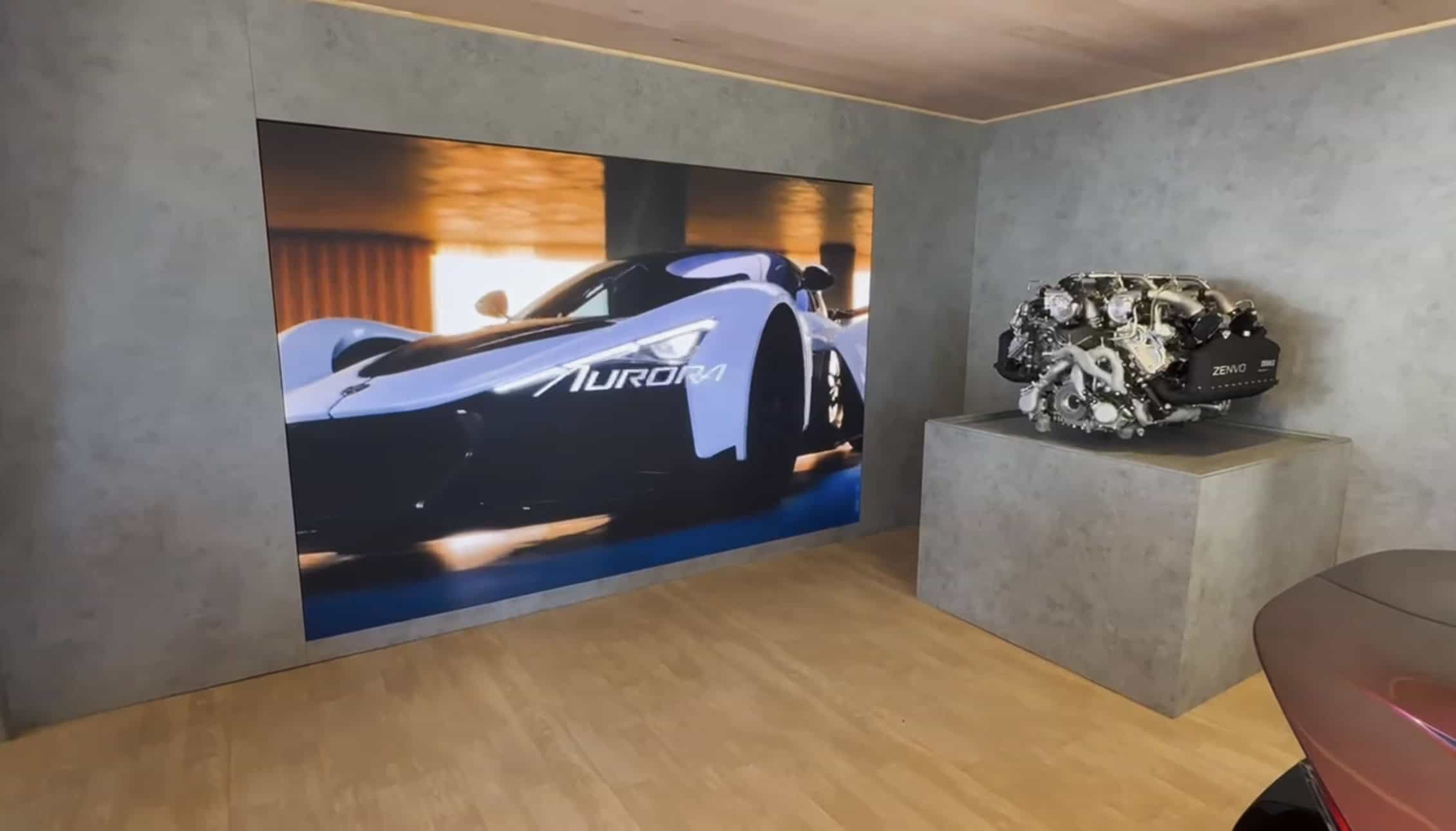Recessed 2mm LED screen in a grey-walled room of the Zenvo pavilion playing a film of the blue Aurora Tur at speed, with the Zenvo V12 engine displayed on a plinth beside it