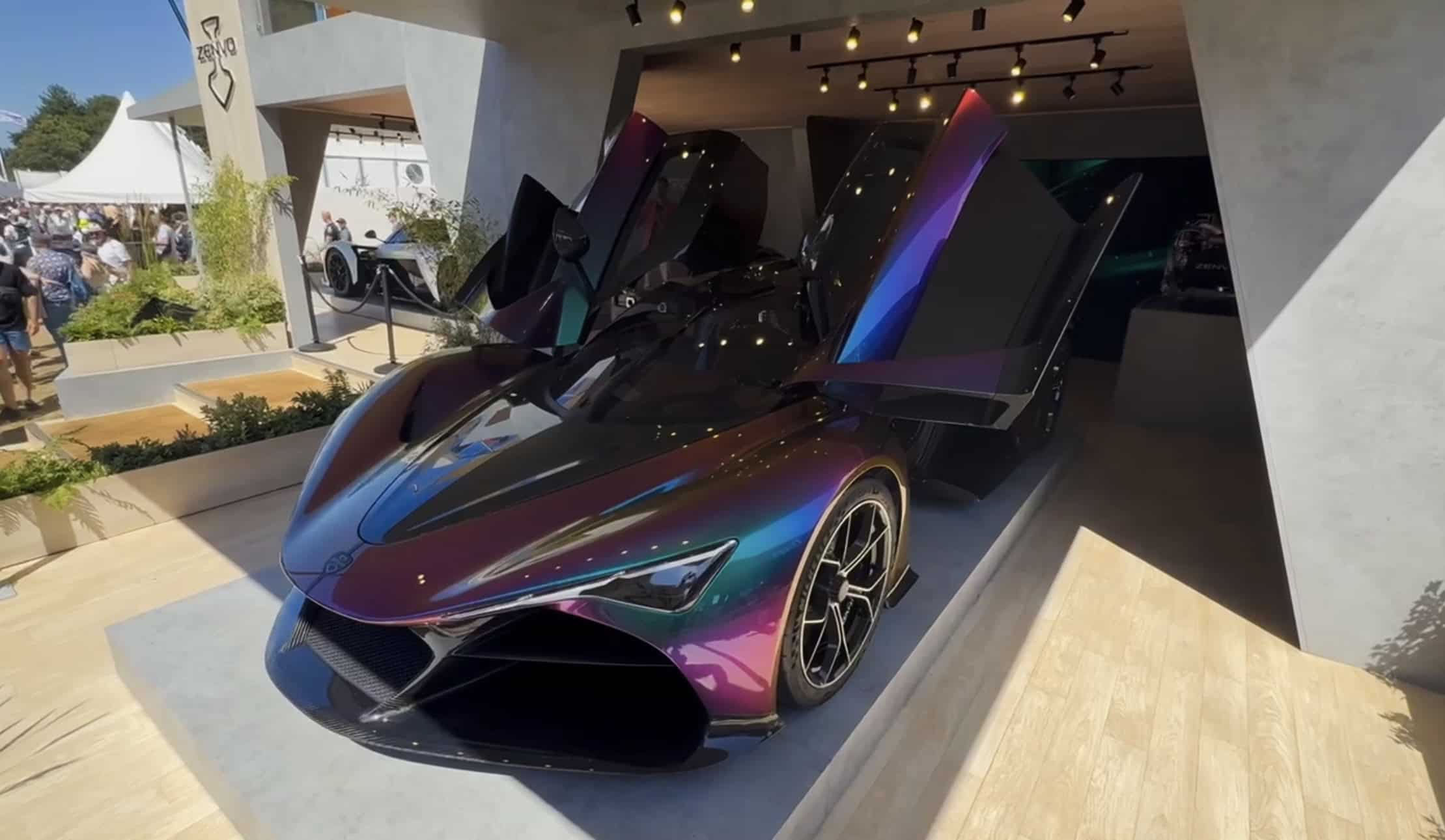 Front three-quarter view of the Zenvo Aurora Tur with both doors raised on a raised timber plinth inside the pavilion, track lighting above and the Goodwood crowd beyond