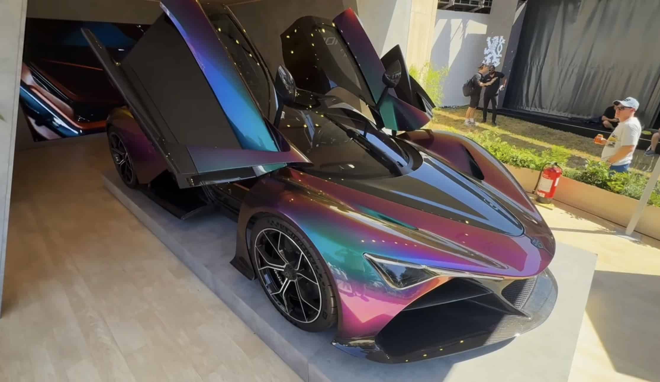Front view of the Zenvo Aurora Tur with its doors raised, iridescent paint shifting from purple to green, with a large LED screen playing car footage visible behind it to the left