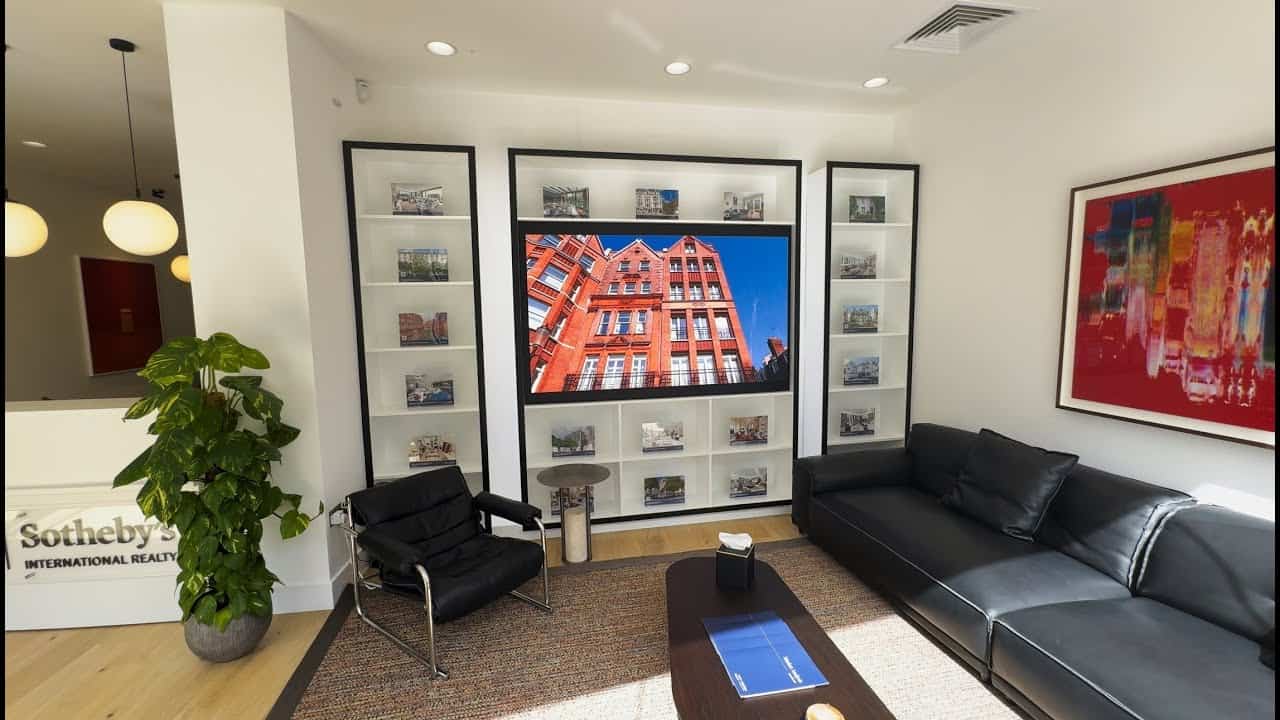 1.25mm DYF Series LED video wall built flush into a white bookcase enclosure in the Sotheby's International Realty office in London, showing a red-brick building against a blue sky above black leather seating