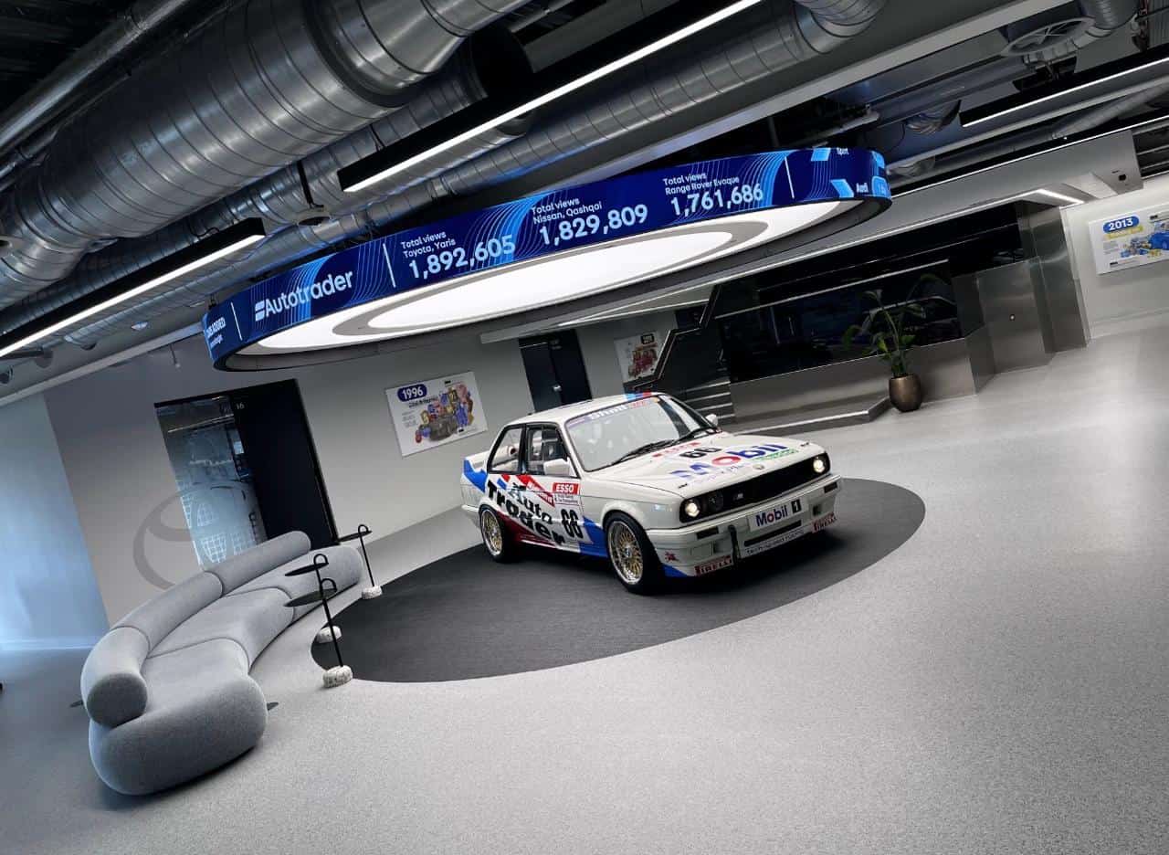 AutoTrader Manchester HQ — circular LED halo ticker showing live vehicle listing statistics suspended above a heritage BMW E30 touring car