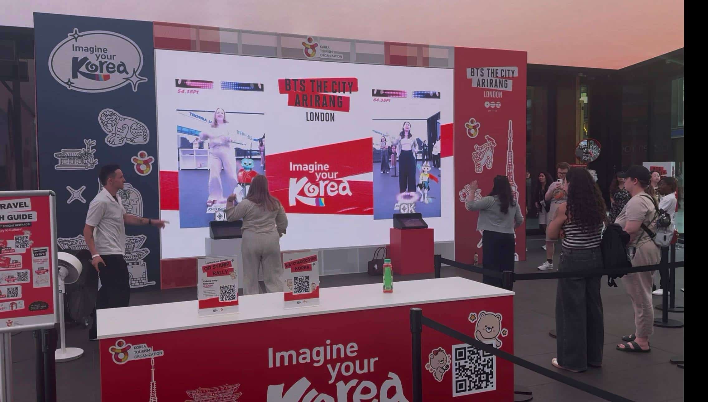 Two visitors playing the K-pop dance challenge at Outernet London, with live camera feeds, animated characters and scores composited on the LED wall in real time