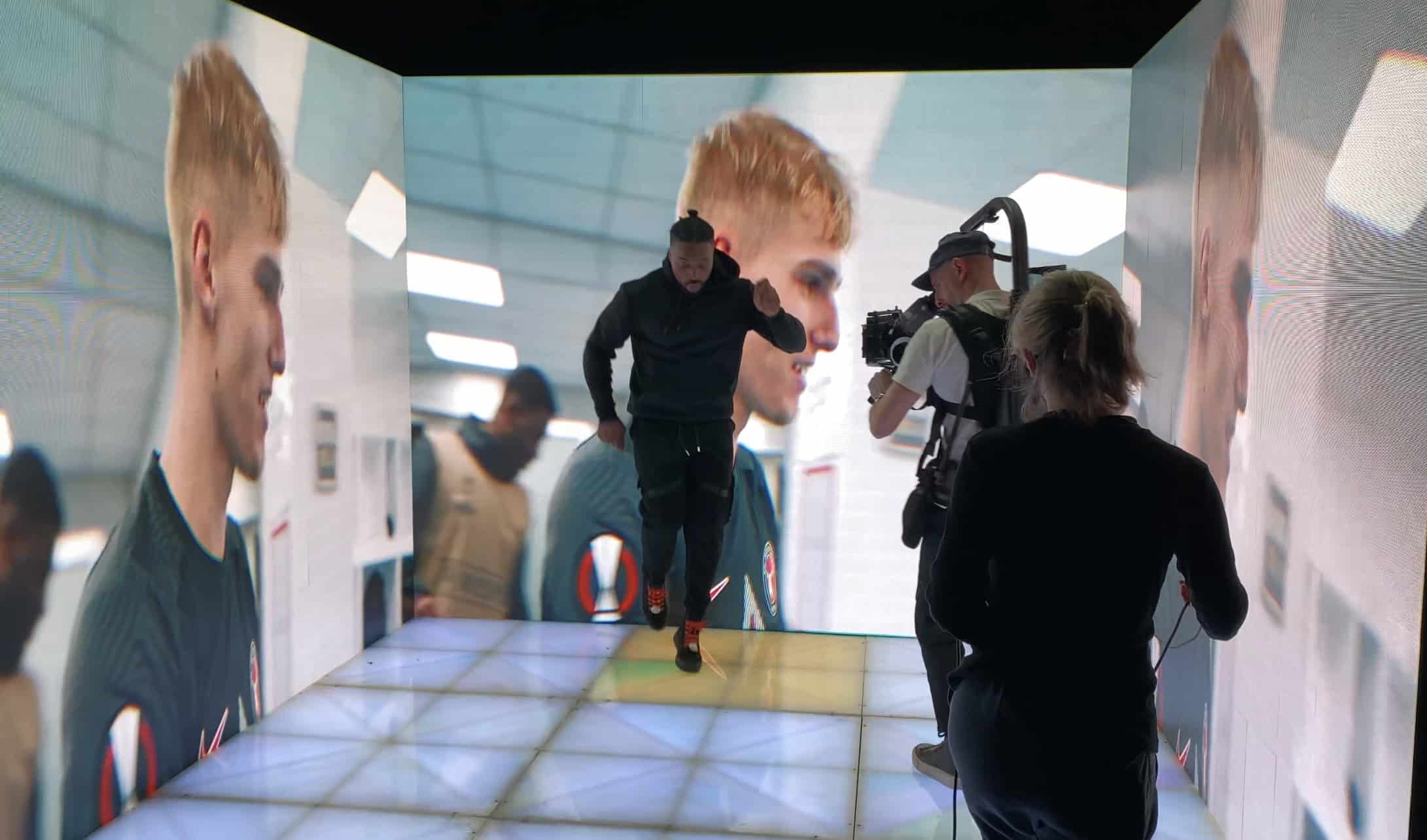 Dancer performing inside a three-sided 2mm LED wall showing close-ups of footballers, a camera operator with a stabiliser rig filming from the open side over a glowing LED floor